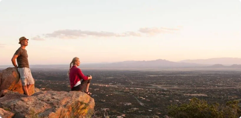 A couple sits on a rocky bluff overlooking a vast, hazy valley and distant mountains at sunset.