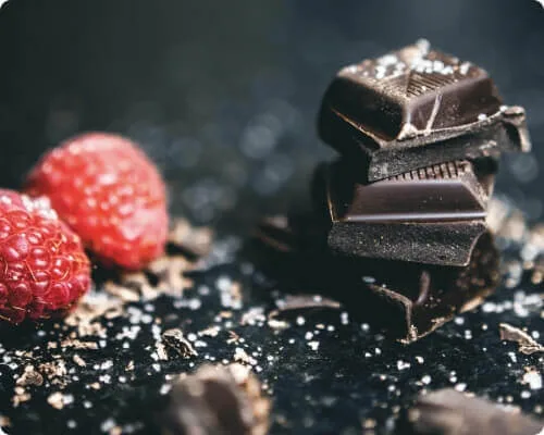 A close-up shot shows a stack of dark chocolate squares next to two bright red raspberries, sprinkled with sugar.