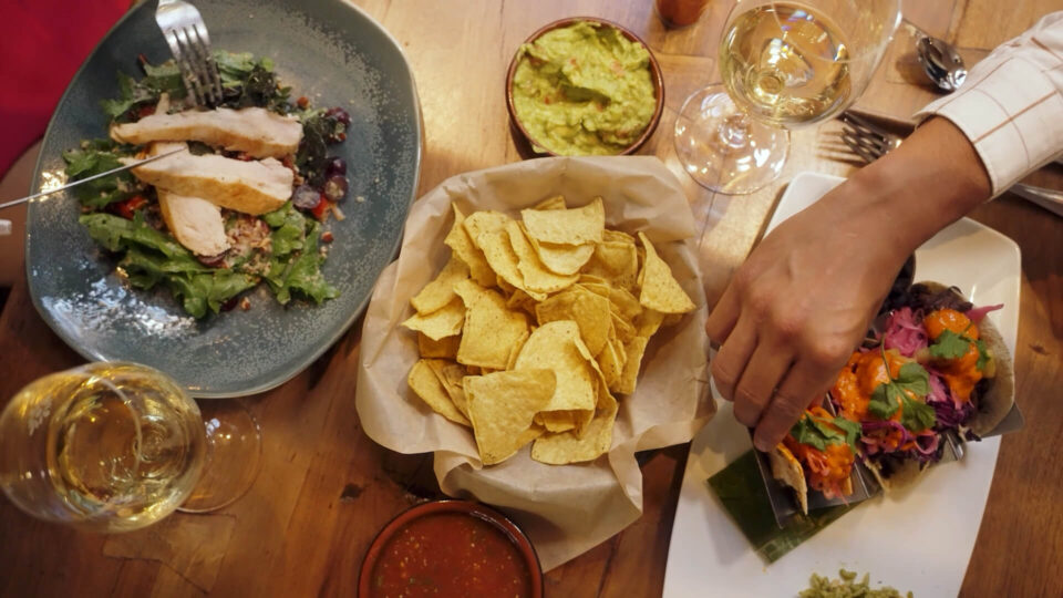 A hand reaches for colorful tacos next to tortilla chips, guacamole, and a fresh chicken salad on a wooden table.