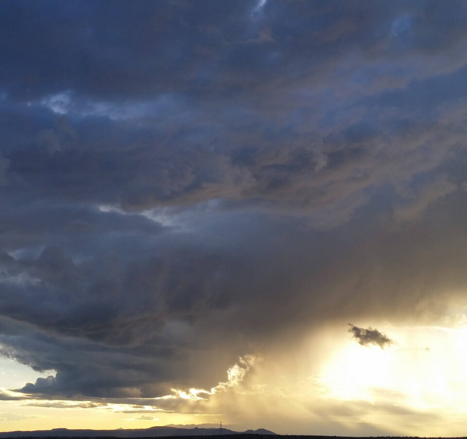 Golden sunlight beams brightly through heavy, dark clouds above a distant mountain range at sunset.