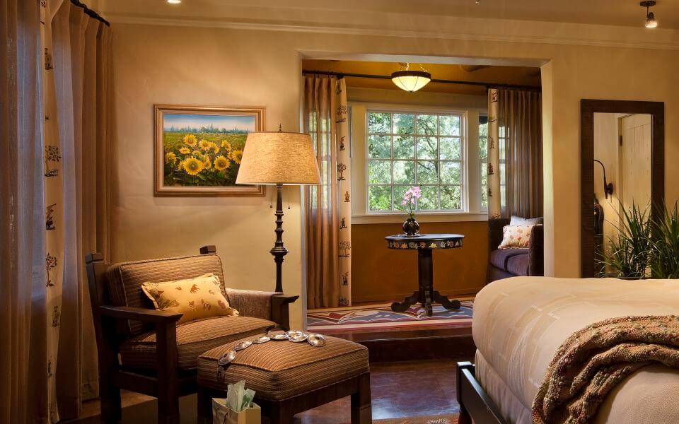 A comfortable, warmly lit boutique hotel room interior with a reading chair, ottoman, and large window.