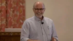 A smiling, balding man wearing glasses and a lavalier microphone speaks passionately during an indoor event.