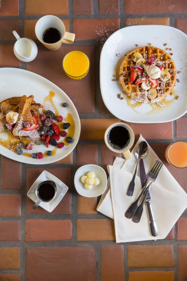 A delicious breakfast setup featuring French toast, a fruit waffle, coffee, and juice served on a tiled table.