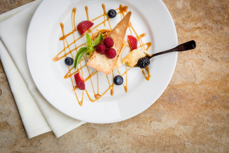 A plated wafer and berry dessert is meticulously drizzled with caramel sauce on a white dinner plate.