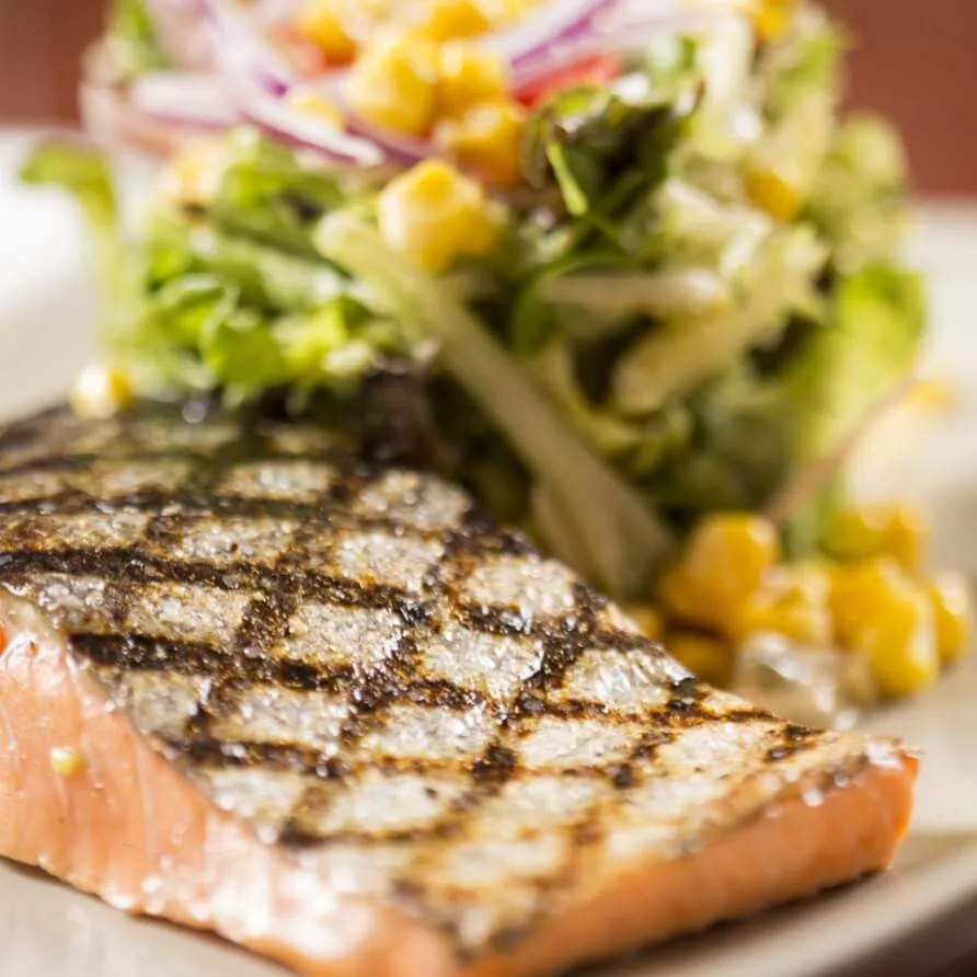 A perfectly grilled salmon fillet with distinct char marks is plated next to a fresh corn and mixed greens salad,.