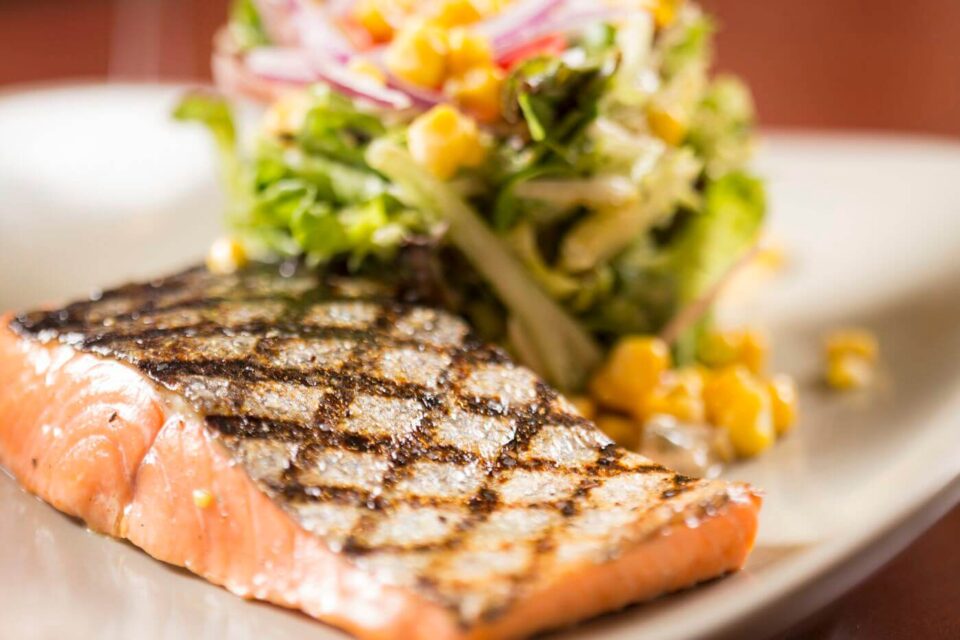 A perfectly grilled salmon fillet with charred grill marks is plated next to a fresh corn and onion salad.