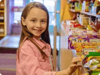 A happy child in a pink jacket reaches for packaged snacks displayed on a shelf in a general store.