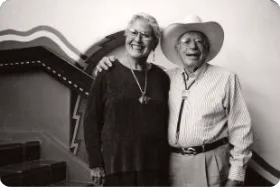 A smiling elderly couple wearing a cowboy hat and bolo tie stands together inside the historic hotel near a decorated.