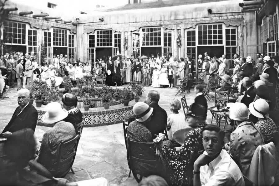 A large group of people in early 20th century attire attend a social event in the beautiful outdoor event space.
