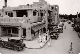 A vintage black and white photograph captures pedestrians and classic cars in front of the historic adobe architecture.
