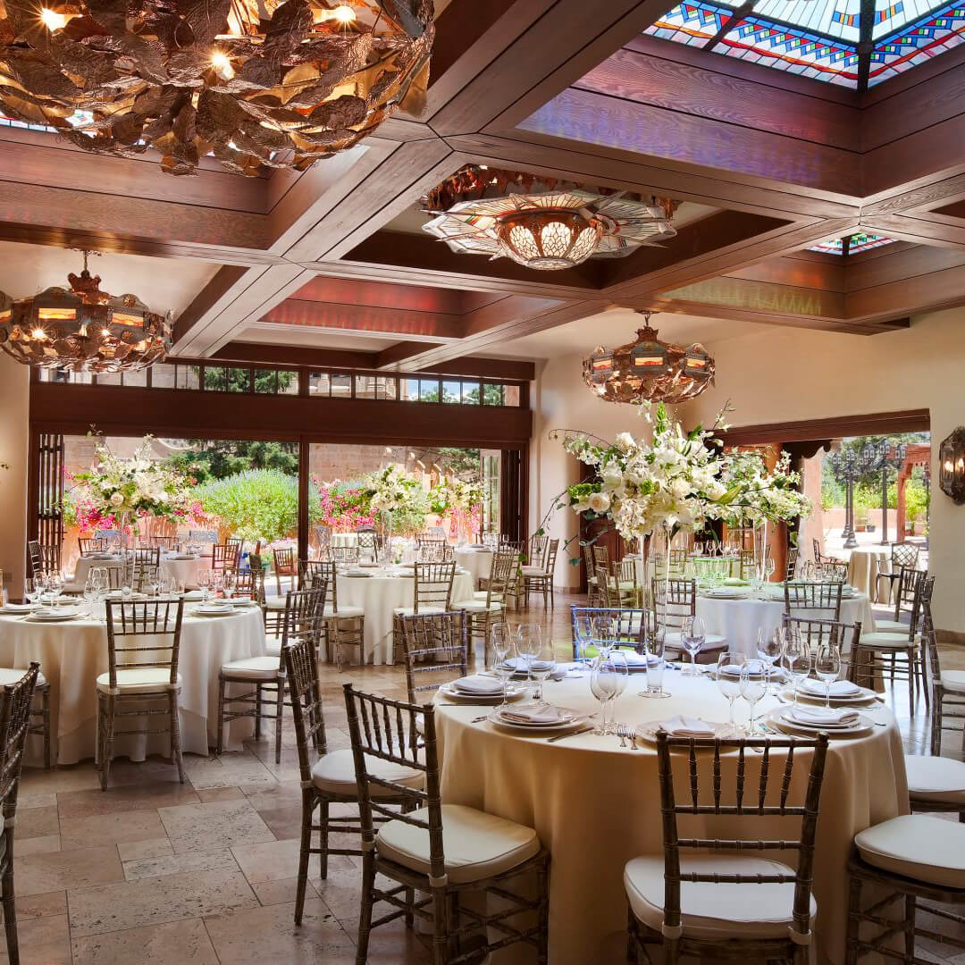 A formal dining room with round tables set for a meal, decorated with flowers and chandeliers.