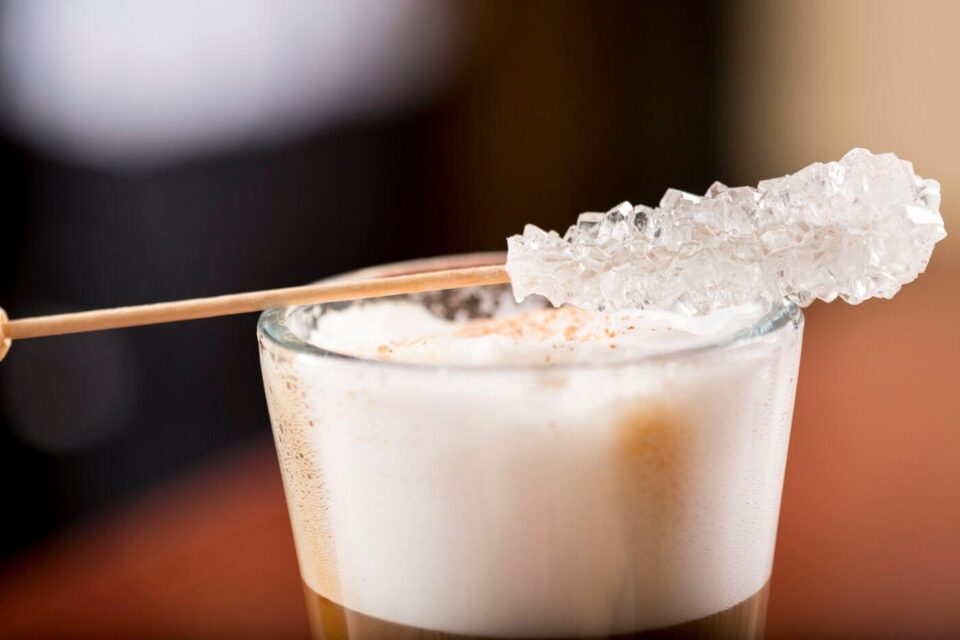 A close-up shows a frothy layered coffee beverage garnished with a long, sparkling sugar crystal stick.