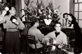 A musician plays guitar and another shakes maracas for guests enjoying live music entertainment.