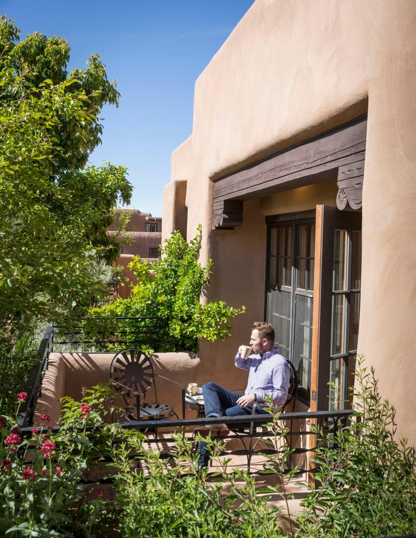 A male guest sips coffee while relaxing on a small, private balcony surrounded by colorful flowers and greenery.