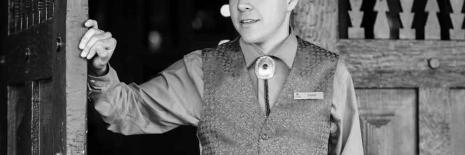 A concierge wearing a patterned vest and bolo tie warmly welcomes guests from a boutique hotel.