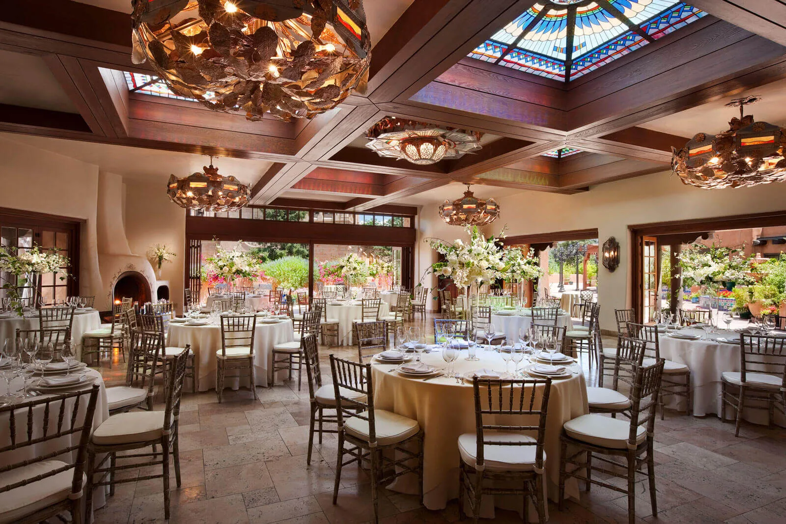 The luxurious ballroom is prepared for a banquet, featuring round tables, large floral arrangements, and a stunning.