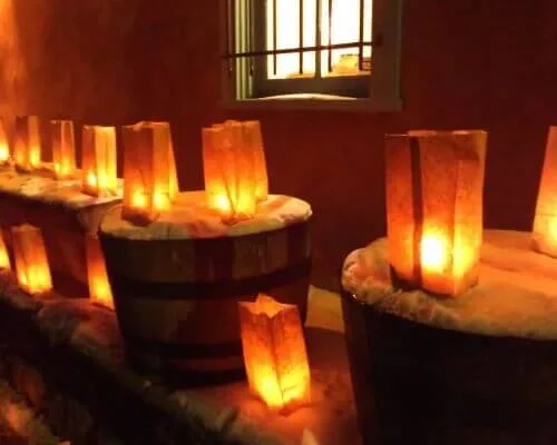 Paper bag luminarias sit on snow-dusted wooden barrels illuminating the adobe entryway at night.