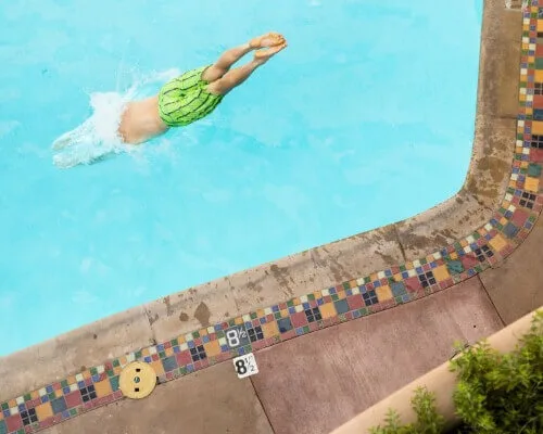 A person wearing bright green trunks dives into the hotel swimming pool, marked by colorful tiled edges.