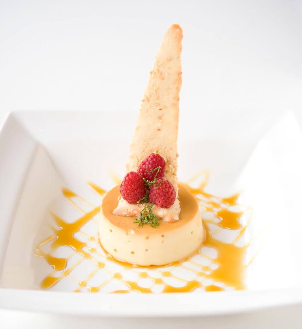 A creamy caramel custard dessert is plated with three fresh raspberries and a tall, triangular cookie garnish.
