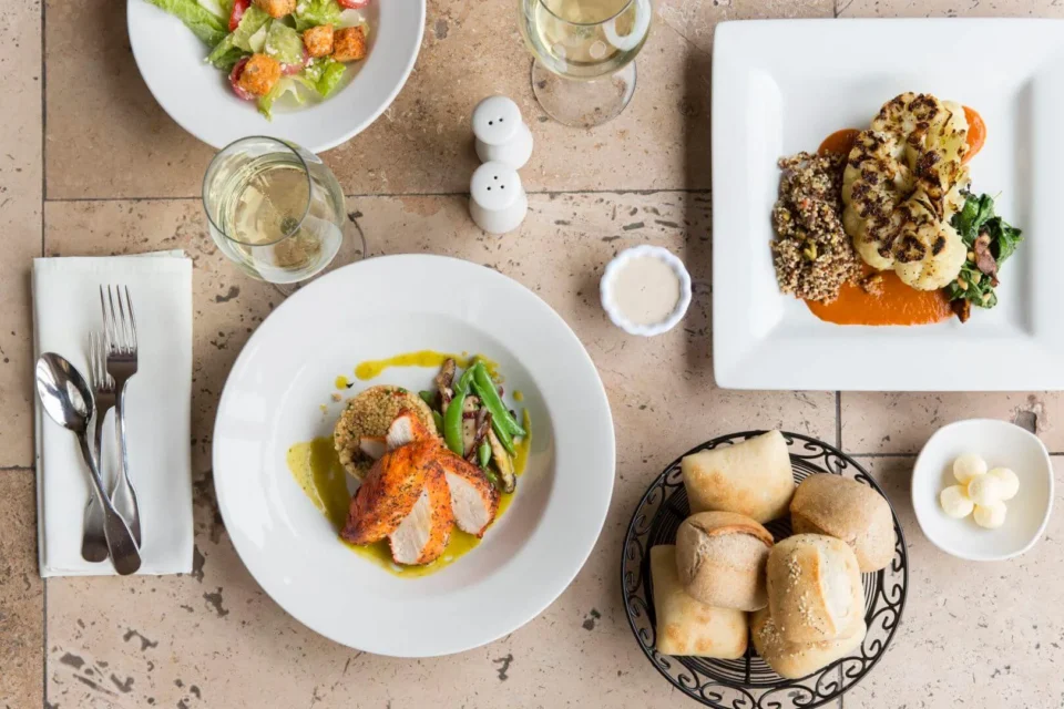 An overhead shot captures two gourmet plated dishes, a side salad, bread, and wine glasses on a tiled table.