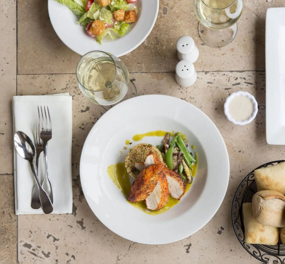 An overhead shot captures a plated chicken entree, fresh salad, and glasses of white wine on a stone dining table.
