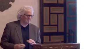 A smiling speaker presents behind a wooden podium in a professional event space.