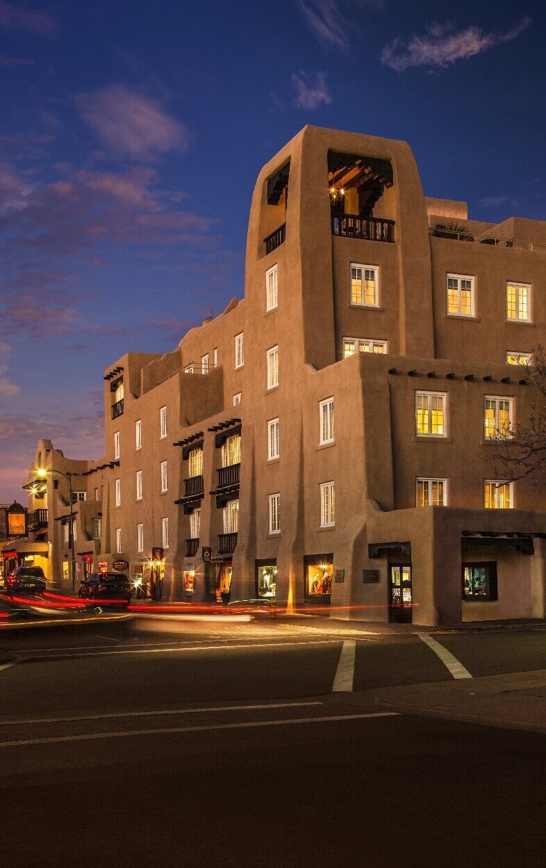 The historic adobe exterior of a downtown Santa Fe hotel glows beautifully against the dark twilight sky.