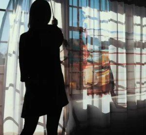 A guest in silhouette pulls sheer curtains, enjoying the bright morning light inside a spacious hotel room.