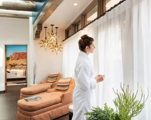 A woman wearing a white robe stands near a window with sheer curtains in the comfortable spa relaxation room.
