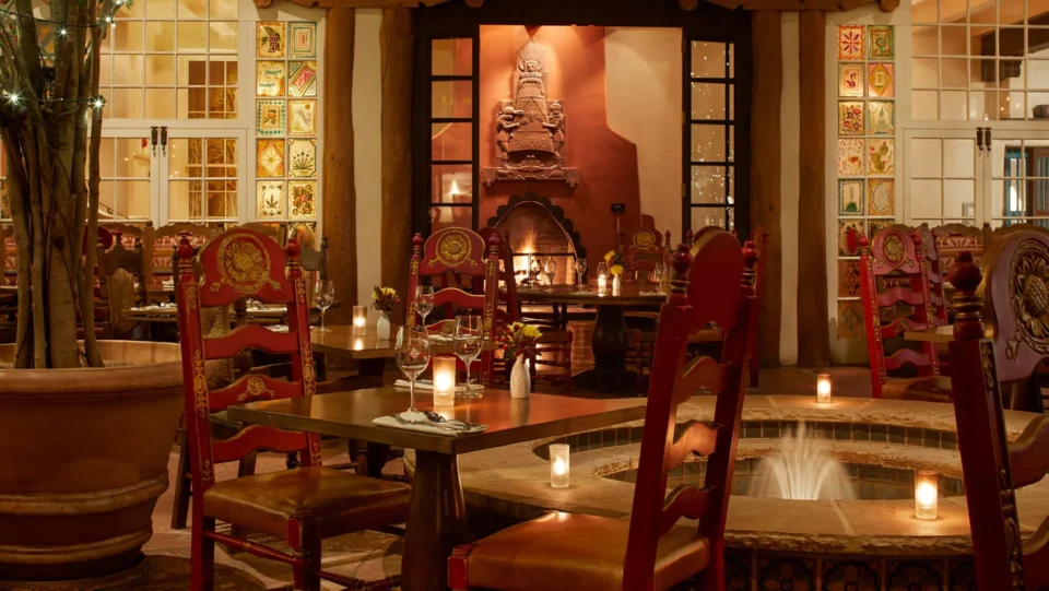 The dimly lit restaurant interior features rustic wooden tables, red Southwestern chairs, a glowing fireplace, and a.
