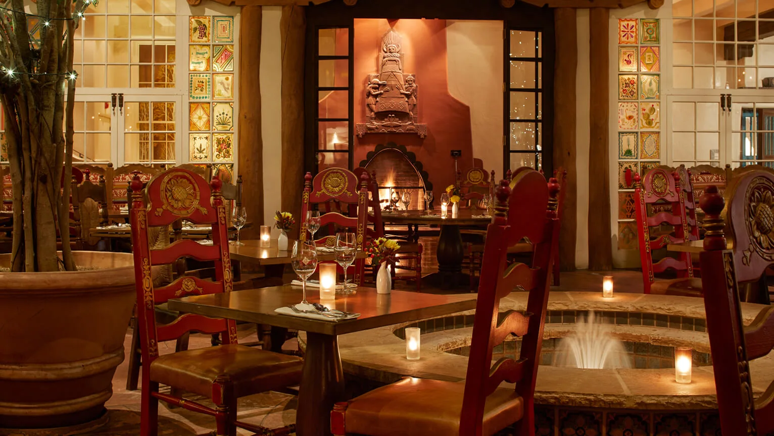 The dimly lit restaurant interior features rustic wooden tables, red Southwestern chairs, a glowing fireplace, and a.