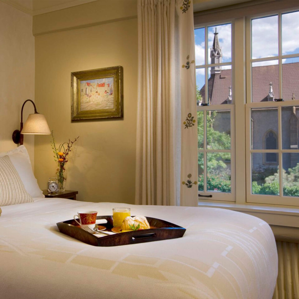 A comfortable hotel room bed holds a wooden tray with breakfast next to a window showing a church steeple.