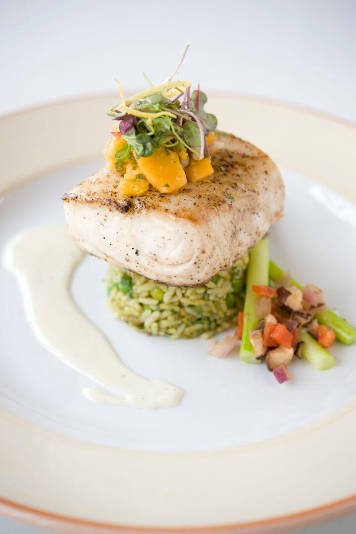 A perfectly plated seared white fish is topped with mango salsa and microgreens, served alongside green herb rice.