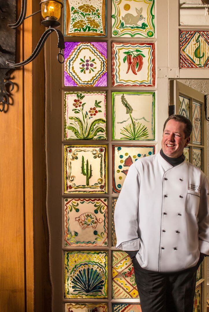 The smiling executive chef stands next to bright, Southwestern decorative tiles in the downtown Santa Fe restaurant.