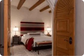 A beautifully carved wooden door opens into a luxurious santa fe hotel room featuring rich textiles and ceiling vigas.