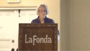 A woman with short gray hair speaks from a branded wooden podium in the hotel's versatile event space.