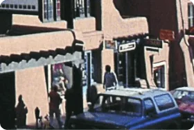 A grainy, historic photo shows a busy street lined with Southwestern adobe buildings, cars, and pedestrians.