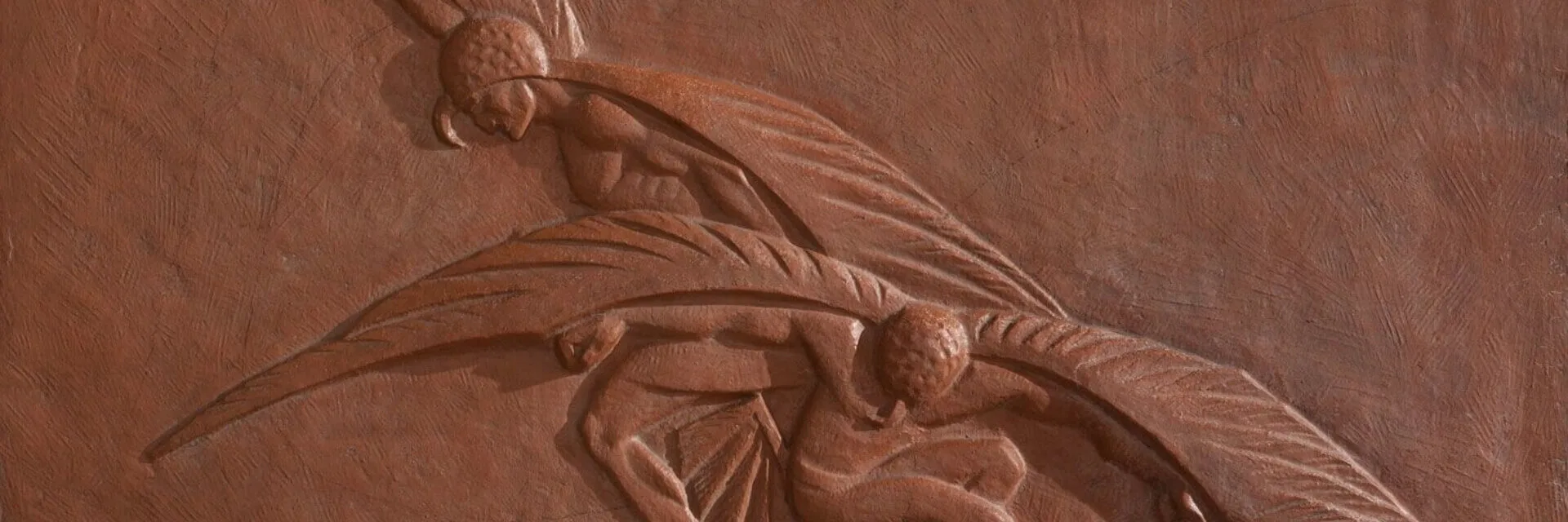 The textured wall features an intricate terracotta relief depicting two stylized, winged figures.
