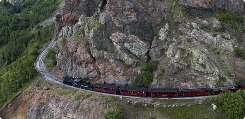 A historic steam train travels along the winding track cut into the green and rocky mountain slope.