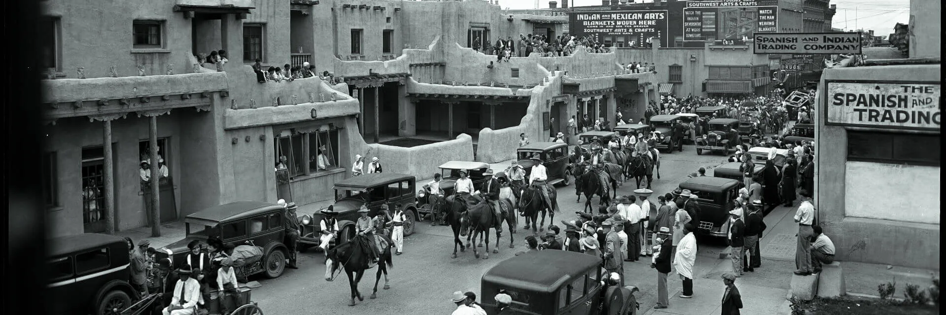 A historic black and white photo shows a crowded adobe street featuring riders on horseback and vintage cars passing by.