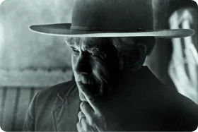 A dramatic black and white image captures a thoughtful older man wearing a wide-brimmed hat and touching his chin.