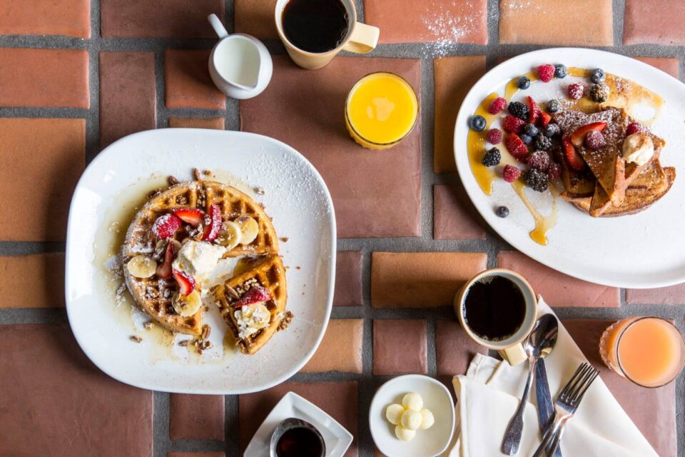Two gourmet breakfast plates, one with a berry-topped waffle and the other with French toast, served with coffee and.