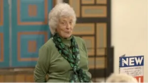 A woman with white hair and a green scarf smiles while standing in front of the patterned wall of an event space.