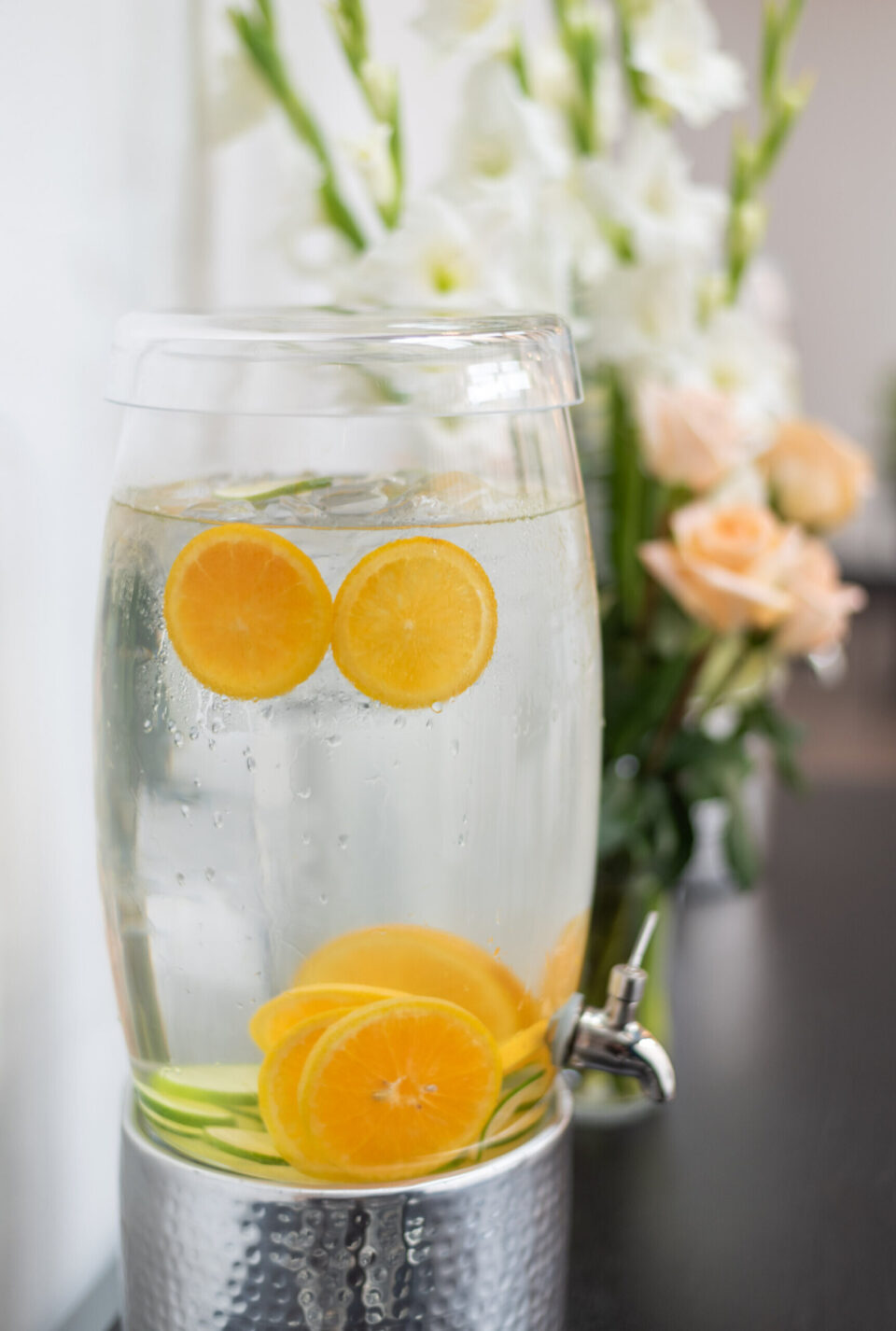 This refreshing citrus-infused water dispenser is set up near beautiful floral arrangements for a banquet hall event.