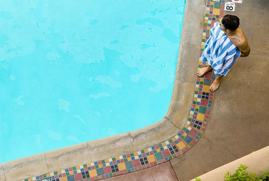 A man wrapped in a striped towel stands barefoot on the brightly tiled edge of the refreshing hotel swimming pool.