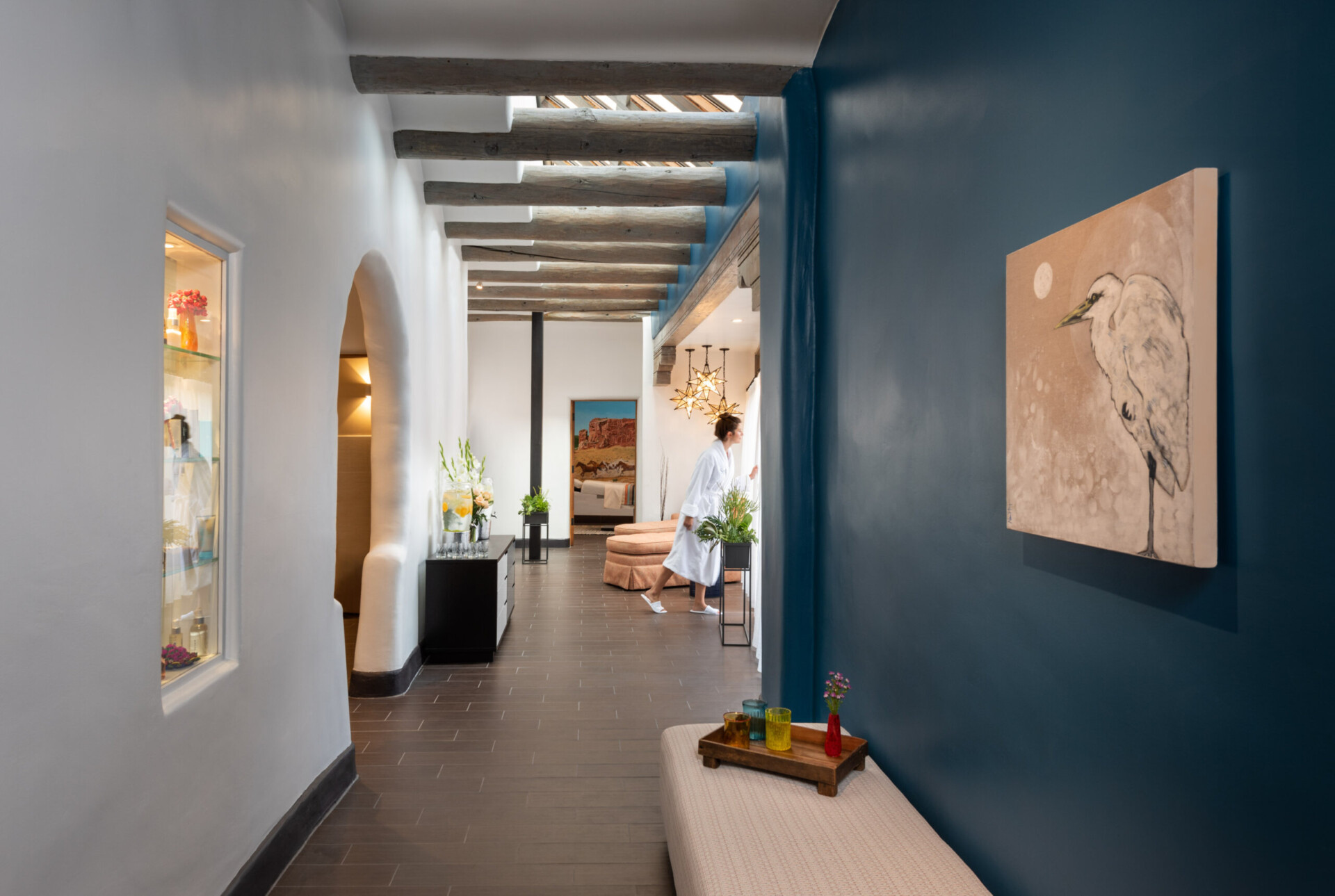 A woman in a white robe stands near the window in the traditional hallway of the spa amenity area.