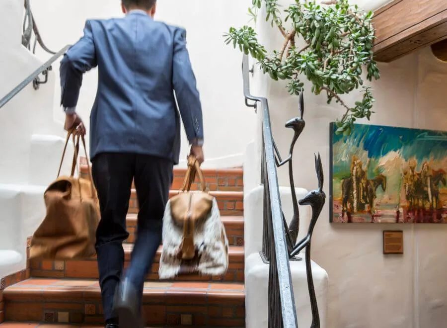 A guest wearing a blue blazer carries two duffel bags up the unique terracotta staircase of a boutique hotel.