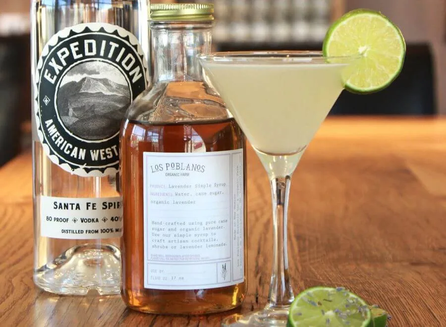 A refreshing lavender martini cocktail is displayed with local spirits and simple syrup ingredients on a wooden bar top.