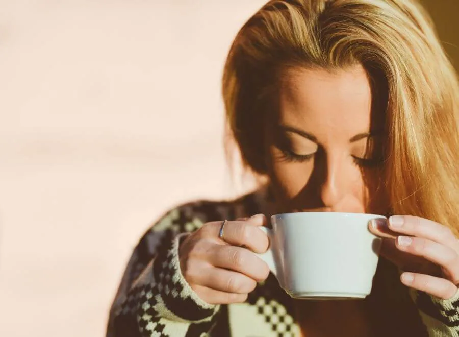 A blonde woman comfortably sips a warm beverage from a large white mug in soft sunlight.