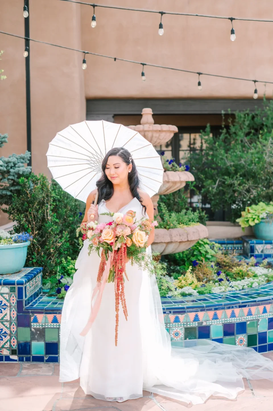 A bride holds a large bouquet and white parasol next to a Spanish-tiled fountain in the wedding courtyard.